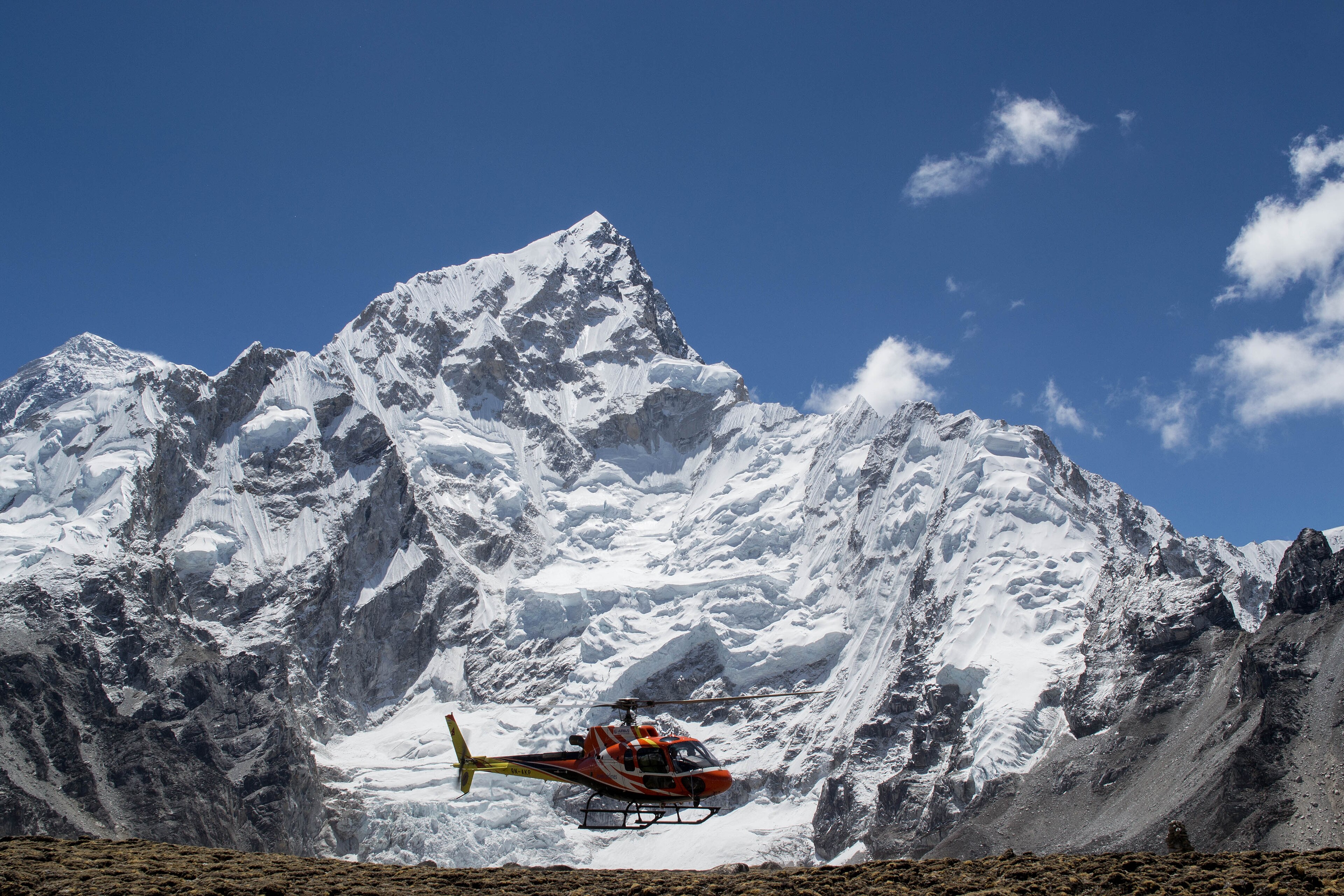 A single-engine helicopter lifting off from a gravel landing site below the Nuptse ice wall, rotors turning.