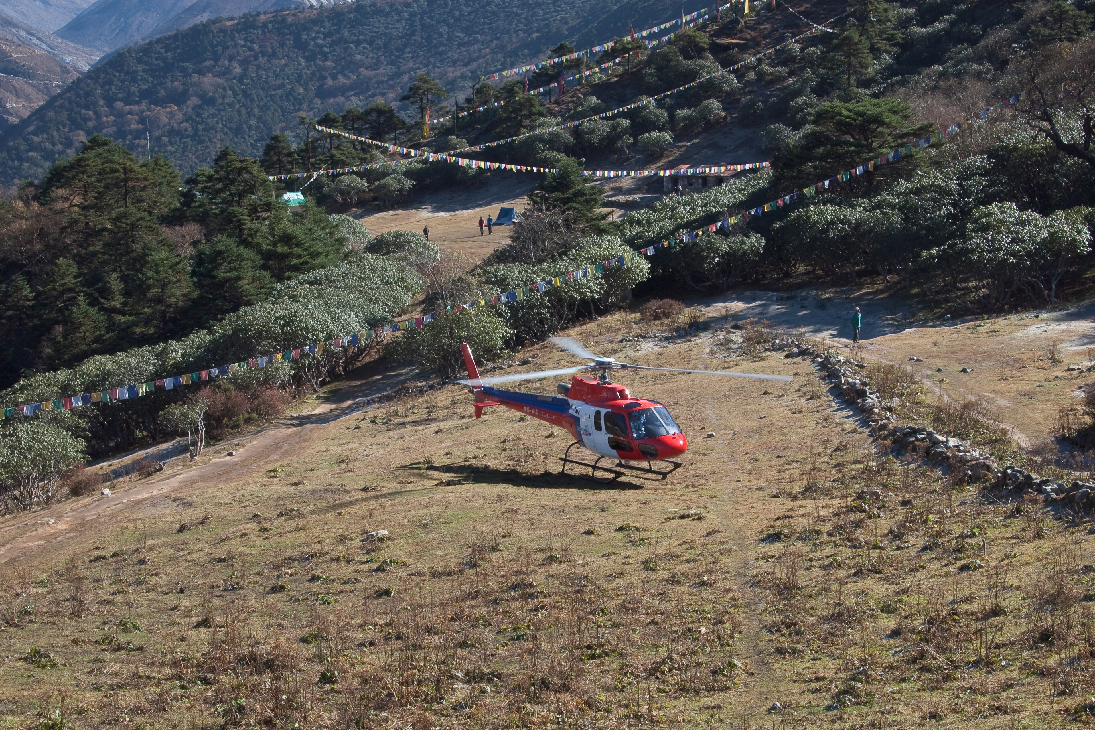A helicopter on a hillside landing ground at Tengboche with its rotor turning, prayer flags strung across the slope.